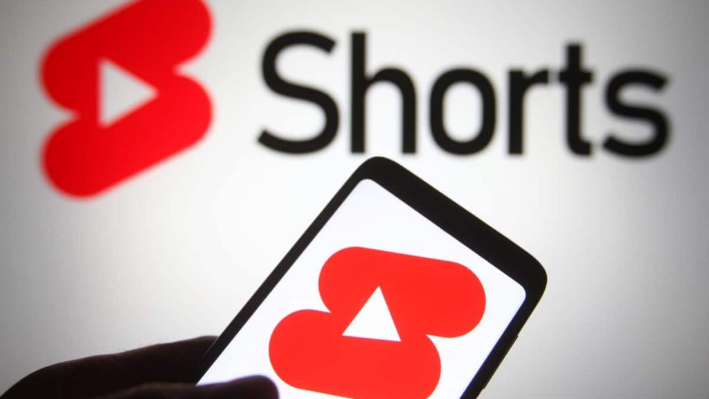 Pessoa segurando celular com logo do YouTube Shorts na tela, também exibido numa tela ao fundo