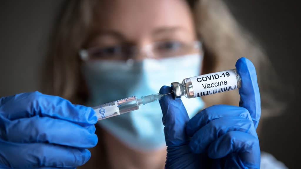 No fundo, desfocado, rosto de mulher com másca e óculos; em destaque, as mãos dela, com luvas azuis, segurando uma ampola e um vidro, no qual está escrito "COVID-19 Vaccine"