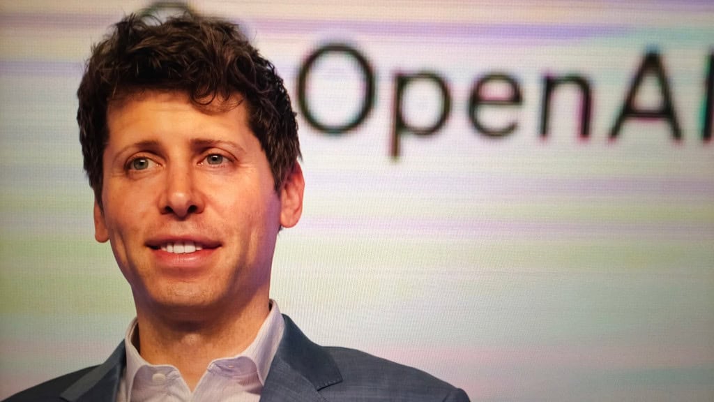 À frente e à esquerda, Sam Altman com um pequeno sorriso; à direita e ao fundo, o logo da OpenAI em uma tela